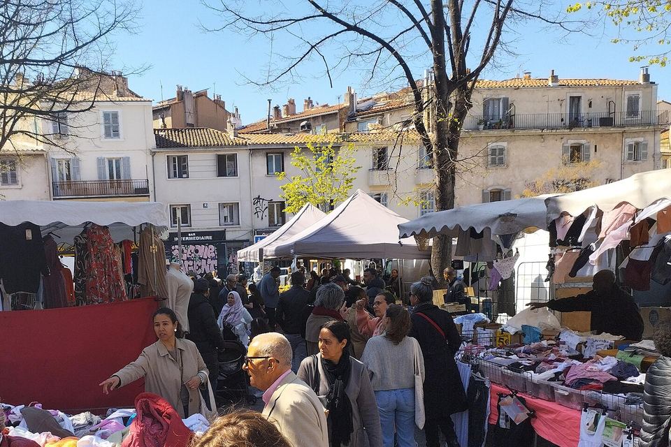 Jour de marché sur une place : étals de vêtements sous des auvents, passants, platanes et façades anciennes.